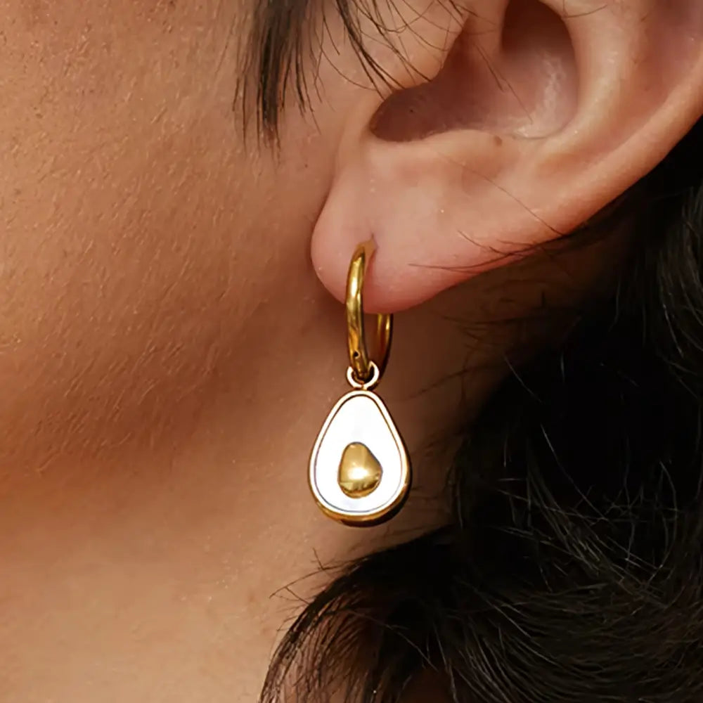 Foto de perto da orelha de da Mulher usando o Brinco Feminino Argola Dourada com Pingente de Abacate de Concha Natural Amara - Banhado a Ouro 18k